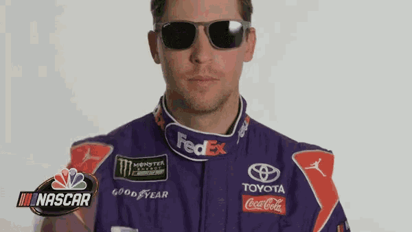 a man wearing sunglasses and a purple race suit is standing in front of a nascar logo