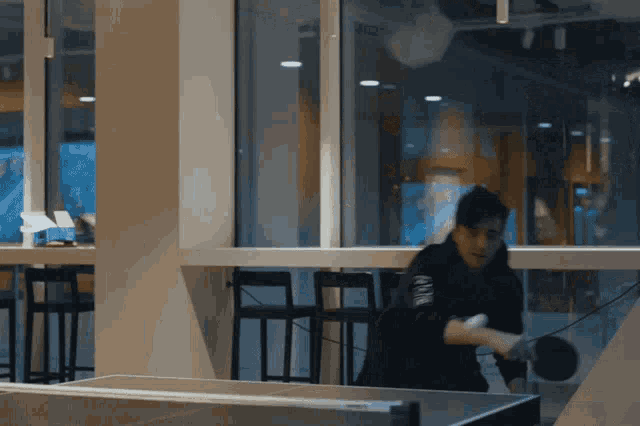 a man is playing ping pong in front of a window with a sticker on his shirt that says ' nike '