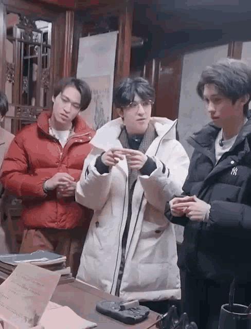 a group of young men are standing around a table . one of the men is wearing a white jacket with the letter ny on it .