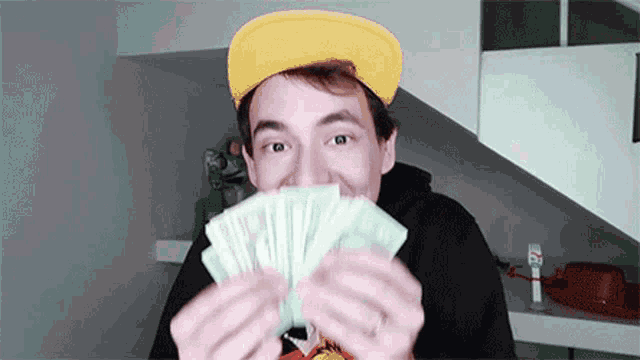 a man in a yellow hat is holding a fan of money in front of his face