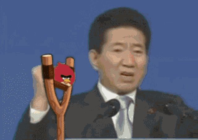 a man in a suit and tie is holding a wooden slingshot in front of a microphone