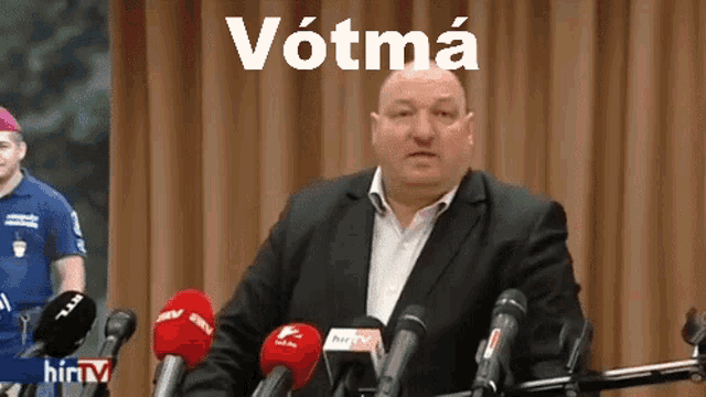 a man in a suit stands in front of microphones with the word votma written above him