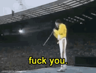 a man in a yellow jacket stands on a stage with a microphone and says " fuck you "