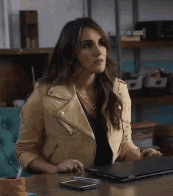 a woman wearing a tan leather jacket sits at a desk with a laptop