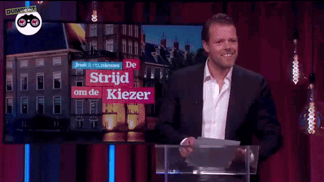 a man in a suit stands at a podium in front of a screen that says " strijd om de kiezer "