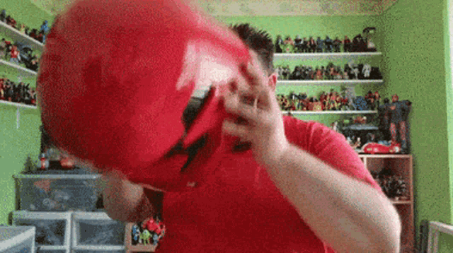 a man in a red shirt is holding a red bucket over his head in a room filled with lots of toys .