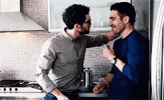 two men are standing next to each other in a kitchen and touching each other 's faces .