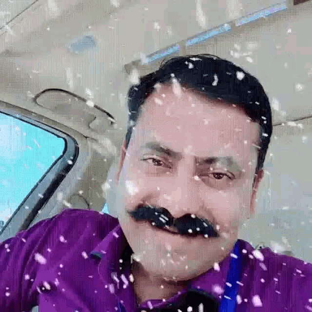 a man with a mustache and a purple shirt is sitting in a car with snow falling on him .