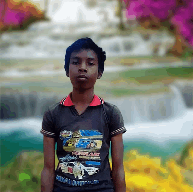 a young boy wearing a ferrari racing shirt stands in front of a waterfall