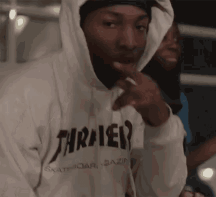 a man wearing a thrasher hoodie is smoking a cigarette and pointing at the camera .