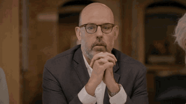 a man with glasses and a beard is sitting at a table with his hands folded
