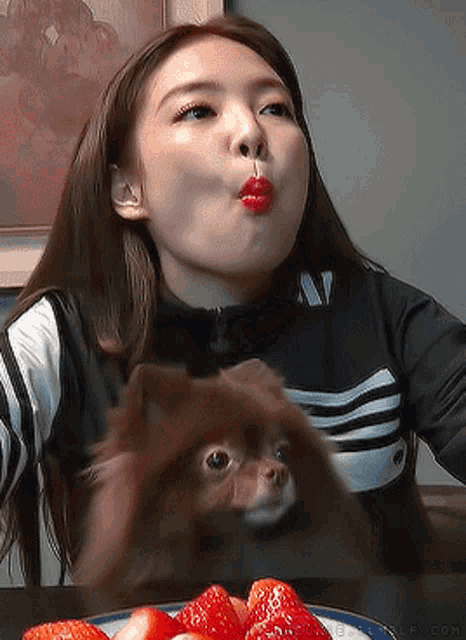 a woman is sitting at a table with strawberries and a dog licking her lips