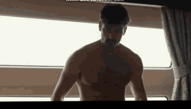 a shirtless man is standing in front of a window and looking out .