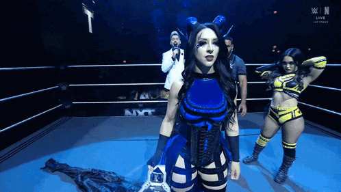 a woman in a blue and black outfit stands in a wrestling ring with the word nxt on the wall behind her