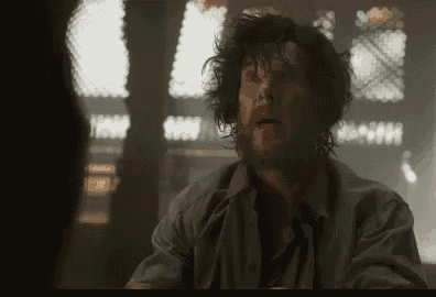 a man with curly hair and a beard is sitting at a table in a dark room talking to another man .