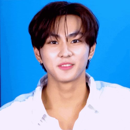 a young man wearing a white shirt and earrings looks at the camera with a blue background