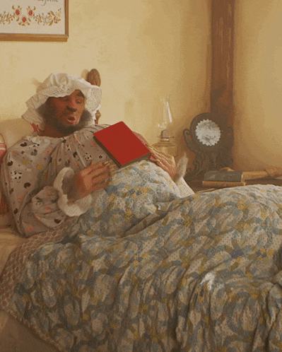 a man laying in bed reading a book with a clock on the wall behind him