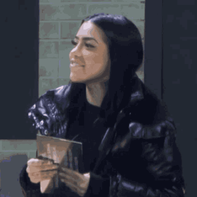 a woman in a black jacket is smiling while holding a piece of paper in her hand .