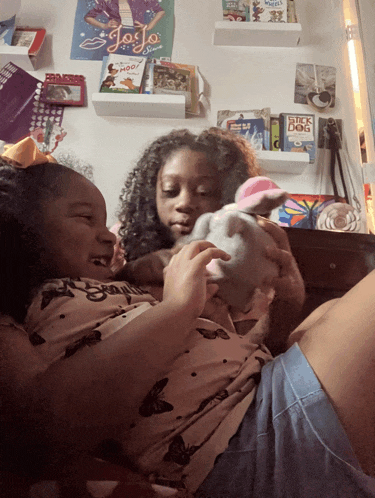 two young girls playing with a stuffed animal in front of a wall with jojo posters on it