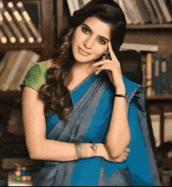a woman in a blue saree is standing in front of a bookshelf with her hand on her forehead .