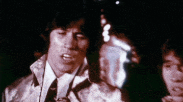 a man in a suit and tie is singing into a microphone while another man looks on .
