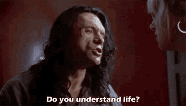 a man with long hair is standing in front of a red wall and says `` do you understand life '' .