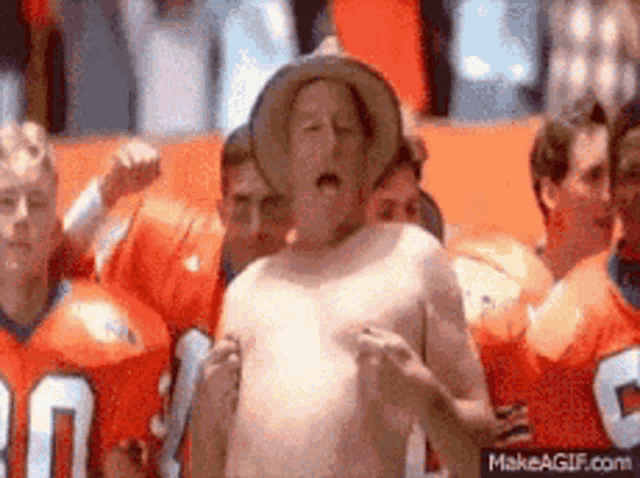 a shirtless man wearing a cowboy hat is standing in front of a group of people wearing orange jerseys .