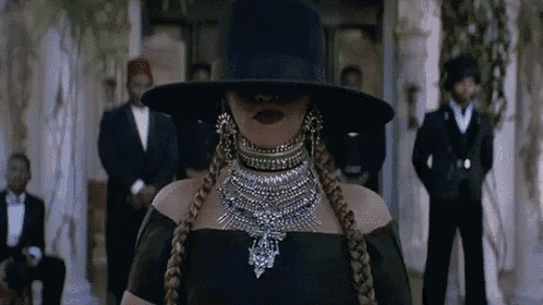 a woman in a black hat and necklace is standing in front of a group of men .