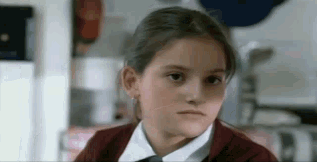 a little girl in a school uniform is looking at the camera .