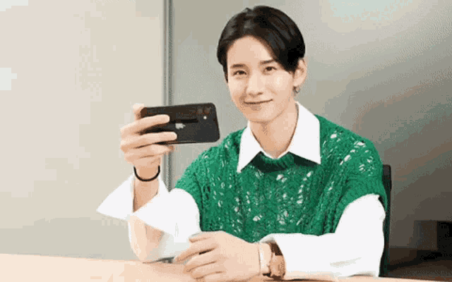 a young man in a green vest is taking a selfie with his cell phone .