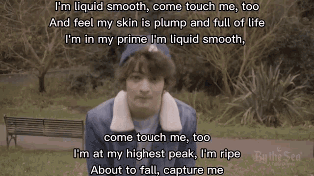 a man in a blue jacket stands in front of a park bench and says " i 'm liquid smooth come touch me "