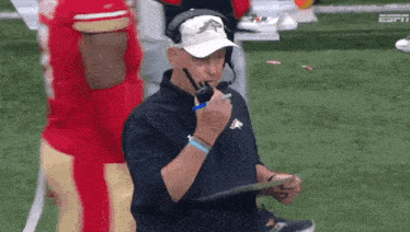 a football coach is talking on a walkie talkie while holding a clipboard on the sidelines .