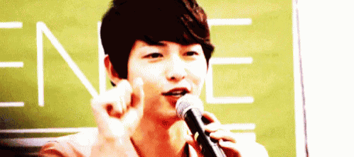 a young man is talking into a microphone and making a heart shape with his fingers .