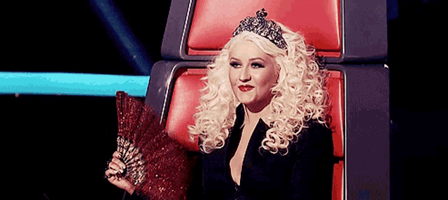 a woman wearing a tiara sits in a red chair holding a fan