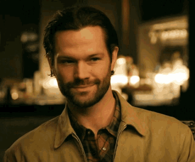 a man wearing a plaid shirt and a tan jacket smiles for the camera