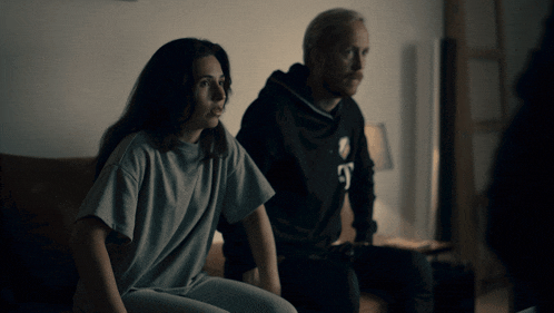 a man and a woman are sitting on a couch and the woman is wearing a grey shirt with the letter e on it