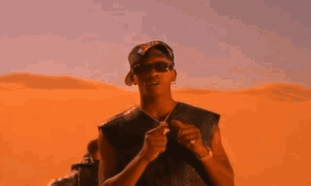 a man wearing sunglasses and a hat is standing in front of a sand dune .
