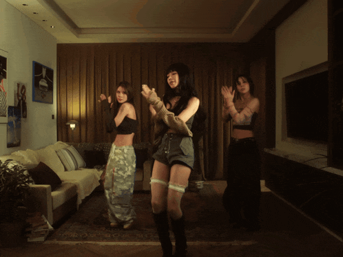 three women are dancing in a living room with a couch and television