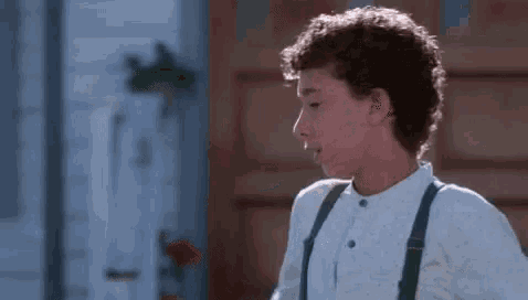 a young boy with curly hair wearing suspenders and a white shirt is standing in a room .