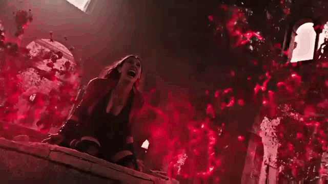 scarlet witch is screaming while sitting on a ledge in a dark room surrounded by red smoke .