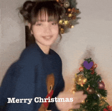 a girl in a blue sweater is standing in front of a christmas tree and says merry christmas