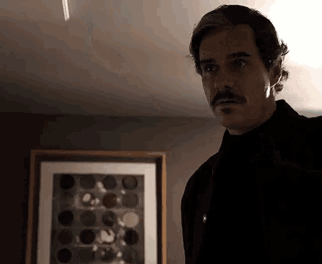 a man with a mustache is standing in a dark room next to a framed picture .