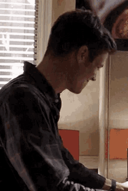 a man in a plaid shirt is sitting at a table in front of a window with blinds .