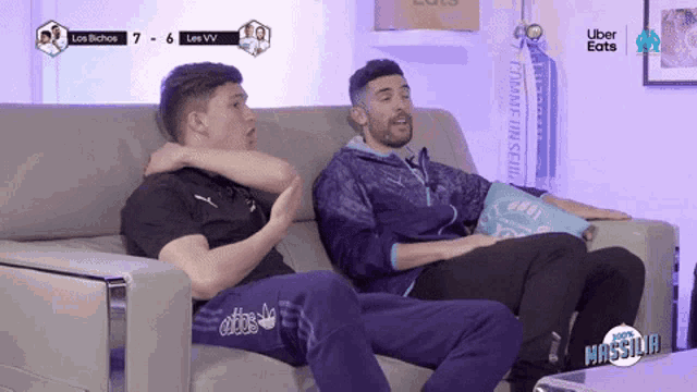 two men are sitting on a couch watching a soccer game with uber eats in the corner