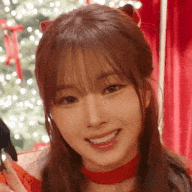 a close up of a woman wearing a red choker and smiling in front of a christmas tree and a christmas tree .