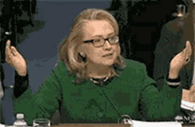 a woman in a green jacket and glasses is sitting at a table with her hands outstretched .