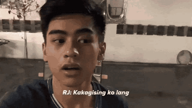 a young man says " rj kakagising ko lang " in front of a white wall