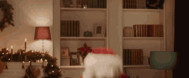 santa claus is standing in a living room in front of a christmas tree and a bookshelf .