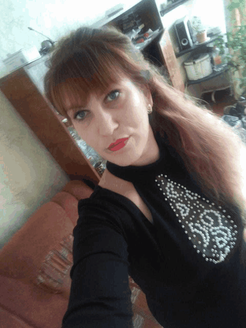 a woman taking a selfie in a living room wearing a black top with pearls on it