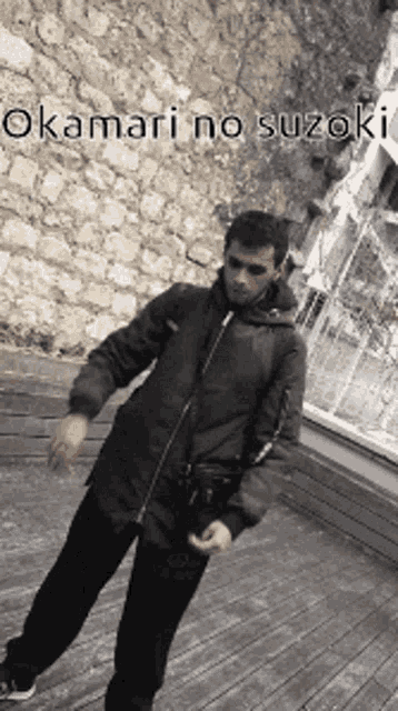 a man in a black jacket is standing on a wooden deck in front of a brick wall .
